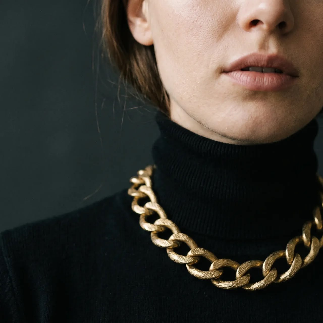 Brushed gold chain collar necklace on a black fine-knit turtleneck, close crop of lips and neck