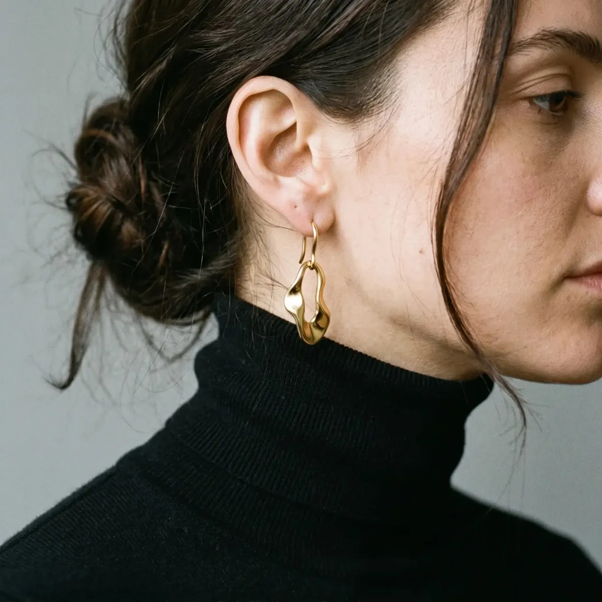 Sculptural gold drop earring against a black fine-knit turtleneck, profile crop with hair in a loose bun